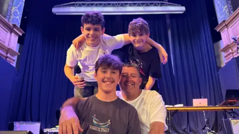 Andrew Turner/BBC Dan and his dad Shaun sitting on the stage. Dan is wearing a brown tee shirt with a logo. Shaun is wearing a white tee shirt, and has his right arm draped over his son's shoulder. Behind them, with arms around each other, are two other band members. They are on the stage in St George's Theatre with curtains behind and a gantry rig above them..