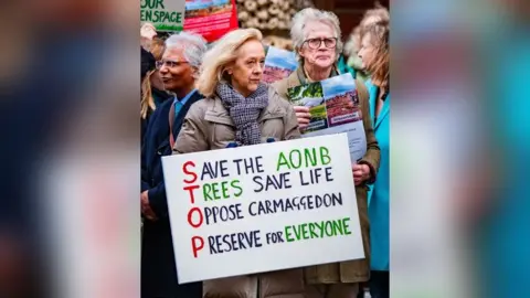 Jamie Bellinger A woman in a coat holding a sign that has 'STOP' written vertically in red letters, with each letter the start of a sentence. The full sign reads 'Save the AONB, Trees save life, Oppose carmaggedon, Preserve for everyone.