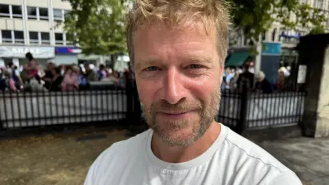 A man with blonde hair and a beard wearing a white t shirt is seen smiling at the camera inside the grounds of Belfast City Hall. Behind him is a black metal fence with crowds gathered behind.