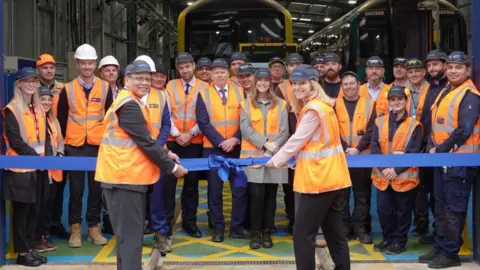 Lucy Rigby MP, on the right, and a male executive from Siemens Mobility, on the left, smile at the camera as they hold a large purple ribbon with a bow in the middle. The ribbon is across the entrance of a large train workshop. The MP and the executive are both wearing orange hi-vis jackets. Behind the ribbon, about 25 employees, also in hi-viz, also smiling, are lined up.