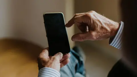 An older gentleman sitting comfortably with his legs crossed, focusing intently on his smartphone. The warm, natural light filters through the room, creating a cozy atmosphere. His hands exhibit signs of age, reflecting experience as he interacts with the device, seemingly absorbed in the digital world. He has on a striped shirt and jeans. The rest of the room is blurred. 