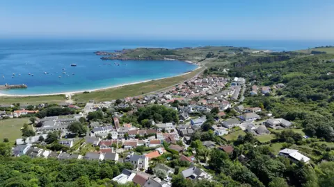 BBC An image taking from above looking down at Alderney. There is a number of houses and trees. There is the coastline in the distance.