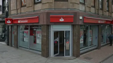 Santander's brach in Whitehaven is on the ground floor of three-storey building on the corner of the High Street. Its entrance door is open. Tha bank's red and white logo reading Santander is placed above the windows. 
