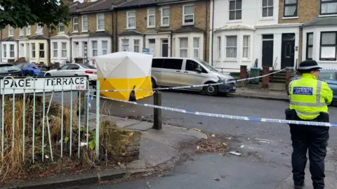 PA Media A Metropolitan Police officer stands near a police cordon and forensic tent on Paget Terrace, near the scene in Eglinton Road, Woolwich, south-east London, where a teenage boy was stabbed to death.