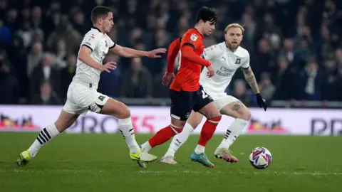 PA Media Luton Town's Liam Walsh (centre) wears red and black and Plymouth Argyle's Jordan Houghton (left) and Tymoteusz Puchacz (right) - wearing white football strips - battle for the ball on the field.