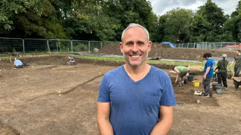 Prof Gabor Thomas smiling at the camera, wearing a blue T-shirt. He has short grey, white hair and facial stubble. In the background people are digging with trowels at the site