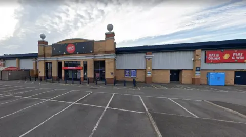 The building is a large warehouse type structure with a large carpark in front of it.  You can "East and drink" and "plan and win" according to the sign.