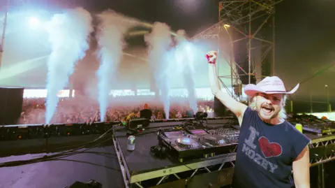 Alastair Brookes A man with I heart Mozey on a capped sleeve navy t shirt. The heart is in diamante detail. They are standing in front of DJ decks and smiling with an open mouth at the camera as they punch their right arm into the air. In the background, you can see what looks like hundreds of revellers and there are steam machines shooting steam into the air. 