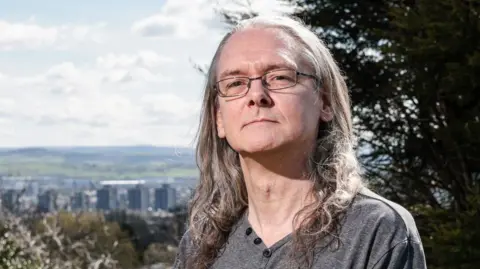 Martin Atkinson, a middle-aged man with long grey hair and glasses, stands looking serious on a grassed area. A cityscape is in the background. 