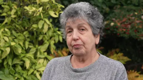 BBC Ruth Jacobs, a woman with grey hair, wearing a grey jumper, is standing outdoors. There is a backdrop of trees.