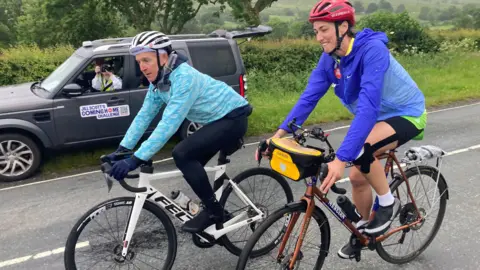 Jill Scott and another bike rider cycle together on a road. There is a support vehicle to the right of them that has a sticker on it that reads 'JILL SCOTT'S COMING HOME CHALLENGE'. It is a rainy day and they are wearing rain jackets.