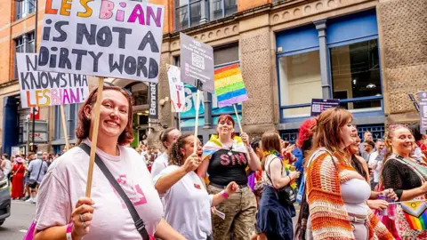 A Pride parade scene in Manchester shows people marching along the street holding signs and rainbow flags. One prominent sign reads “Lesbian is not a dirty word”. Colourful outfits and banners fill the lively crowd against a backdrop of brick buildings.