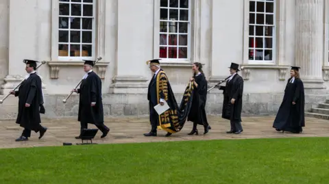 University of Cambridge A row of college officials, dressed in black mortar boards and gowns, walk in a line across the side of a university building. Three of the seven people are carrying maces. Lord Smith is wearing a gown with a train, which is being carried by a woman behind him.