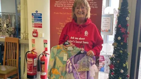 Revive Centre Charity A woman in a red jumper, wearing glasses, and holding two dressing up outfits 