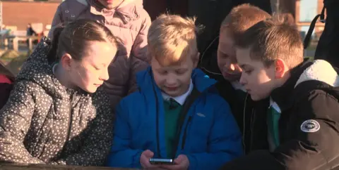 Six children dressed in winter coats huddling around a phone displaying one of the stories