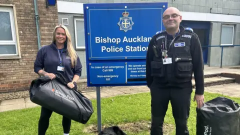 A man and a woman, smiling, stand outside a police station next to a blue sign which reads 'Bishop Auckland Police Station'.
To the left, a woman with long blonde hair, wearing a dark blue hoodie, holds a big black duffel bag with the words 'Sleep Pod'; to the right, stands a man wearing glasses and a police uniform - there is a Sleep Pod, also in a duffel bag, on the ground beside him.