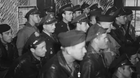 IWM Duxford A black and white image showing at least 18 US pilots in 1943 at Duxford. They mostly standing in rows, sideways on and looking intently forward. They are all wearing US uniform, some with caps and some with peaked hats. 