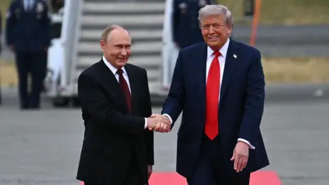 Getty Images Trump and Putin, both smiling, facing the camera and in dark suits, shake hands on the red carpet on the Anchorage airstrip tarmac. Stairs to a plane, flanked by people in uniform, are seen out of focus in the background. 