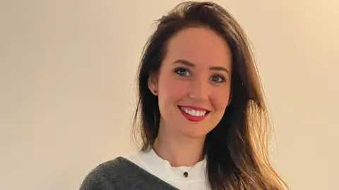 A head and shoulders shot of a woman with long brown hair, wearing red lipstick. She is smiling in front of a white backdrop. She is wearing a white tshirt with grey v-neck jumper and a short chain necklace with green gemstone.