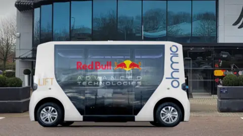 Smart City Consultants A driverless bus parked on a forecourt. It is a shuttle and is white. It has both Red Bull and Ohmio branding on it