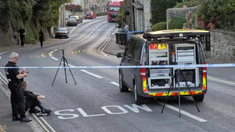 BBC A police van on the main road in Woodstock. Blue tape surrounds the scene, with some numbered markers in the road. Police officers stand by.