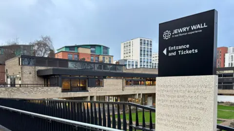 A sign for the Jewry Wall museum with the museum itself in the background