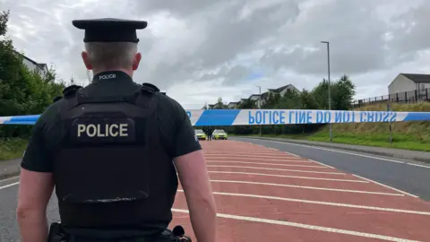 A police officer in short sleeves and wearing a cap stands facing the police cordon