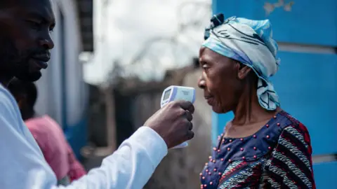 AFP via Getty Images A man in a white shirt holds a contactless infrared thermometer close to a woman's face. She is looking away from the camera and is wearing a blue headscarf and a patterned red, blue and white dress with red beads on it.