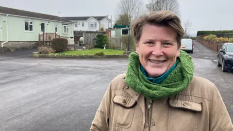 Sarah Dyke is standing in a car park in front of some houses. She is wearing a brown jacket and a green scarf. She is smiling.