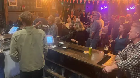 Claire and Neal Hensley Lots of people in a crowded pub. The view is from behind the bar looking onto the clientele. There is bunting with the Union Jack on and a dartboard on the far wall