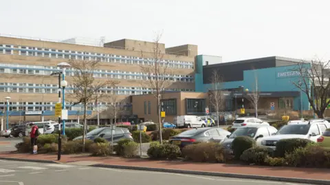 STSFT Sunderland Royal Hospital is a five-storey brick building with rows of windows. A section of the building, which is blue, juts out. There is also a car park out front.