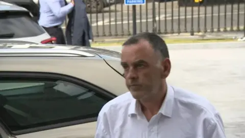 RTÉ  Tim Gilchrist in a white shirt pictured entering court with a silver car in the background