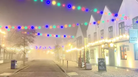 Snaphappydave/Weather Watchers Colourful Christmas lights can be seen strung across a road between buildings in Kinver with other lights attached to apex-roofed buildings