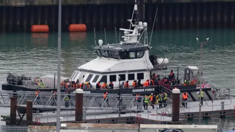 A Border Force vessel is at a harbour on 1 April 2026 with a group of people wearing life jackets standing on deck and along the gangway, with crew members also visible and port buildings in the background.