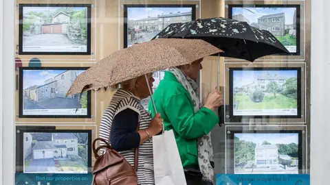 Two people carrying umbrellas walk past an estate agents window displaying houses for sale, in Holmfirth on 29 September 2022