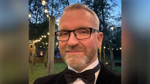 Contributed Mr Lait smiles at the camera while wearing a black suit with a bow tie and white shirt. He appears to be at a party or function. It is getting dark and there are strings of lights between poles behind him.