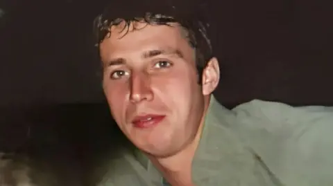 A man with dark hair and brown eyes wearing a green shirt looks at the camera