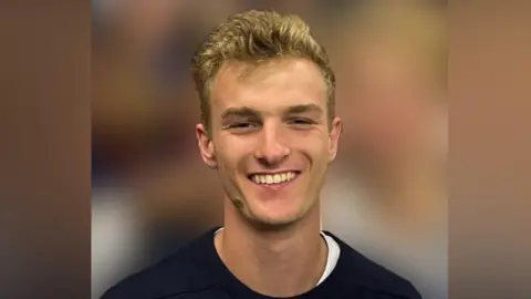 Michael Henry smiles as he looks directly into the camera. He has blonde hair, brushed back from his face. He is wearing a dark sweater.