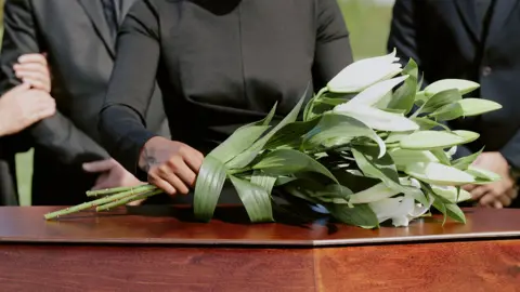 Stock photo shows white flowers being placed on a wooden coffin by people attending a funeral outdoors.