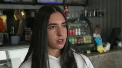 A young woman stands in front of a counter. She has long, straight black hair and wears a white hoodie.