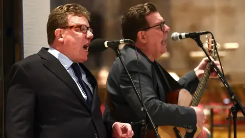 PA Media The Proclaimers, twins with dark hair, dark glasses and dark suits, sing into microphones alongside each other on stage. 