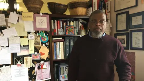 The World Curtiss Reed Jr is shown standing in a room with many documents posted on the wall.