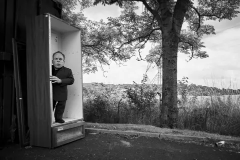Charlie Gray Warwick Davis stands in an empty wardrobe and looks at the camera