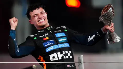 Lando Norris is holding up a trophy while celebrating his win of the Formula 1 championship. He is wearing his kit and is smiling while closing his eyes and pumping his right fist in the air.