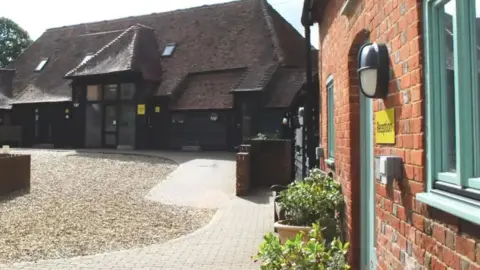 A dark farm-style building with tall and wide glass doors at the entrance sits at the end of a gravel path, with a red-brick building on the left-hand side which has a green windowsills and a green door and a yellow sign on the brick wall reads "reception"