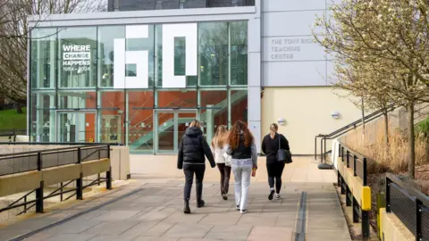 Four young women have their backs turned walking towards a modern university building. They are wearing jumpers and coats. There are a line of trees to the right. The floor is paved with concrete slabs.