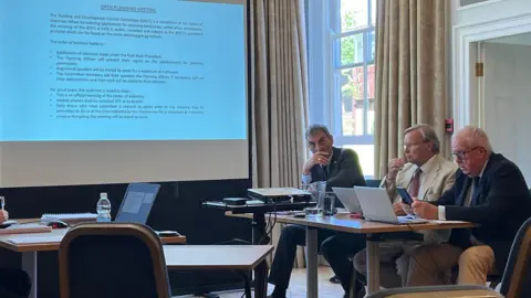 Three men sit in a meeting room, with laptops in front of them and a projector.
