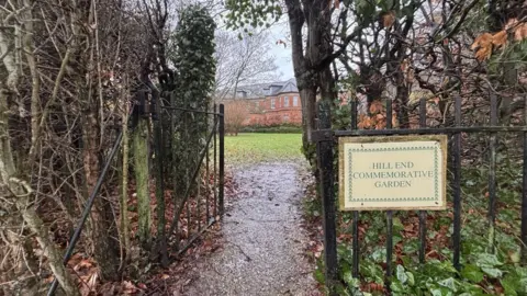 Katy Lewis/BBC An open metal gate leading to a park with a red brick building in the background. There is a sign next to the gate which reads "Hill End Commemorative Garden".
