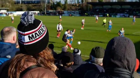 Getty Images A crowd watch a football match. The players are out of focus in the background But you can see the backs of the crowds heads. They are mostly wearing coats and hats. The woman closest to the camera has long brown hair and is wearing a black and white bobble hat with the word Maidenhead on the side.