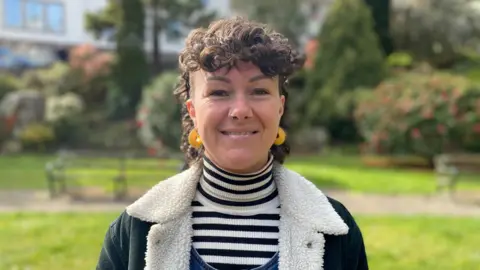 Deputy Bury has dark brown hair. She is wearing a black and white striped turtle neck t-shirt with a dark blue, white fleece lined jacket.